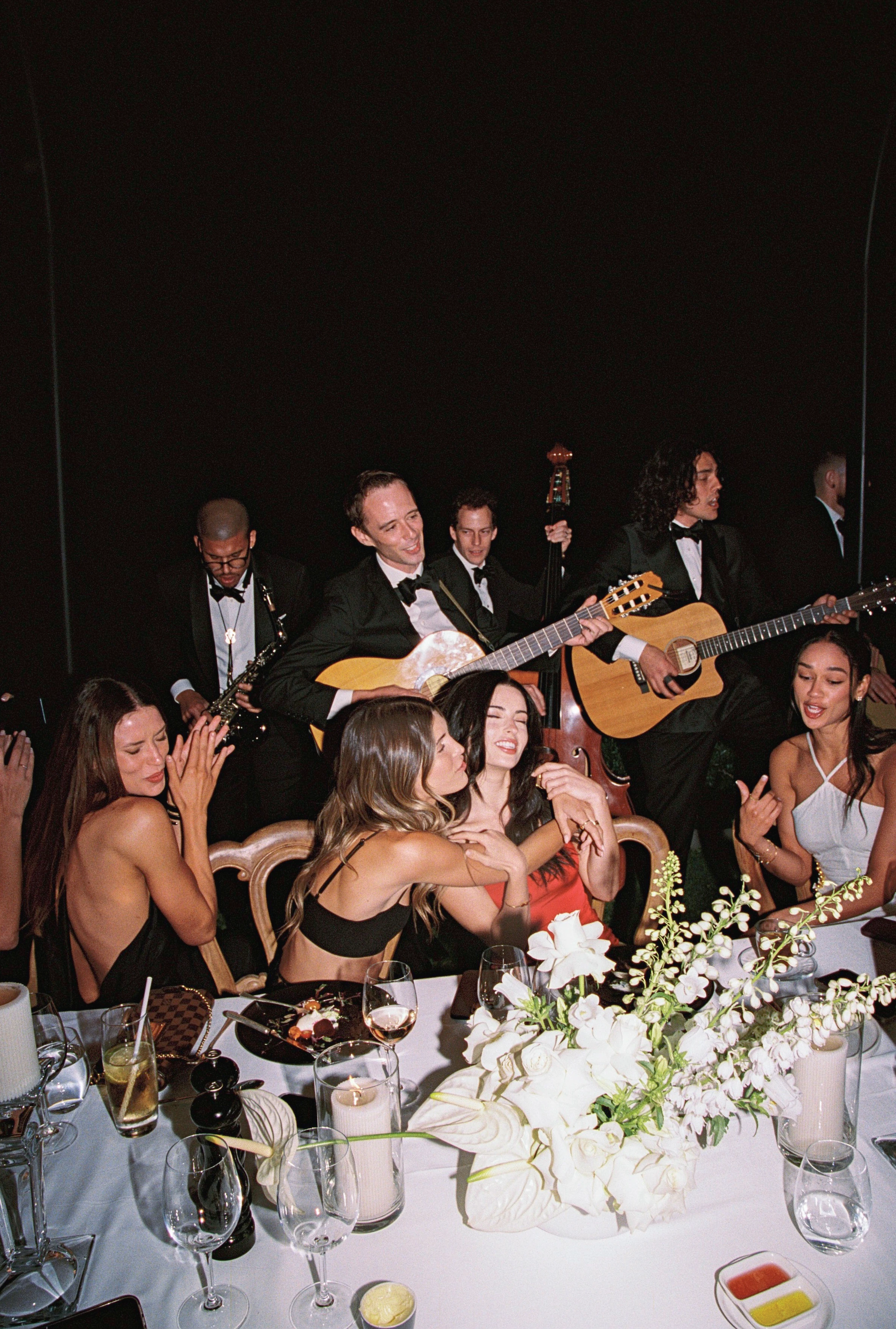 Empire musicians in black tie playing saxophone, guitars and double bass among guests singing around a dinner table
