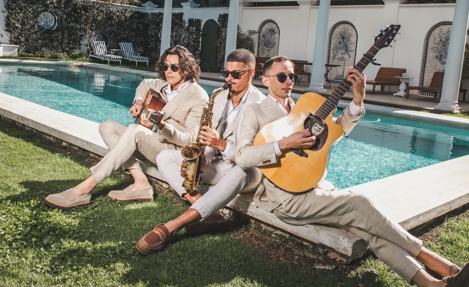 Two Empire guitarists and a saxophonist performing beside a swimming pool in light suits